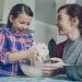 Little pretty girl helping her mother in the kitchen stirring dough for cookies into bowl. Mom and daughter have fun talking and laughing. Family, food, home and people concept